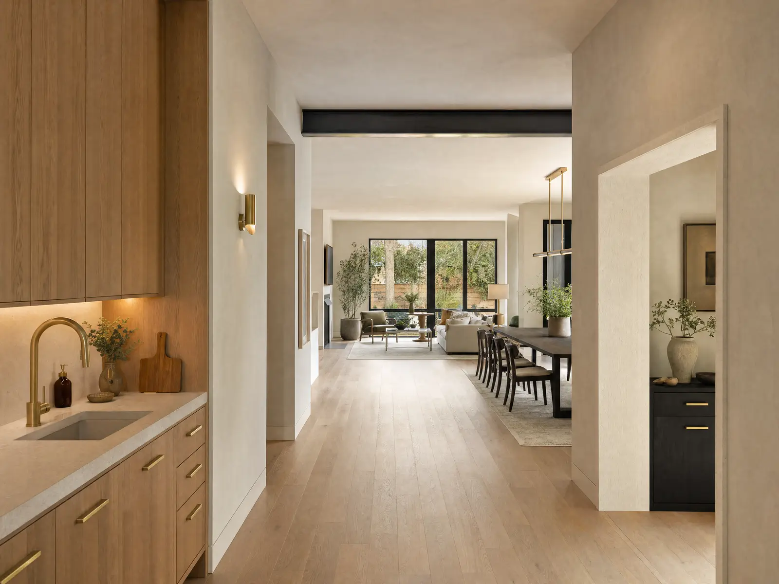 A hallway looking through a wide cased opening into an open living and dining space, with continuous oak flooring and a steel beam line in the ceiling.