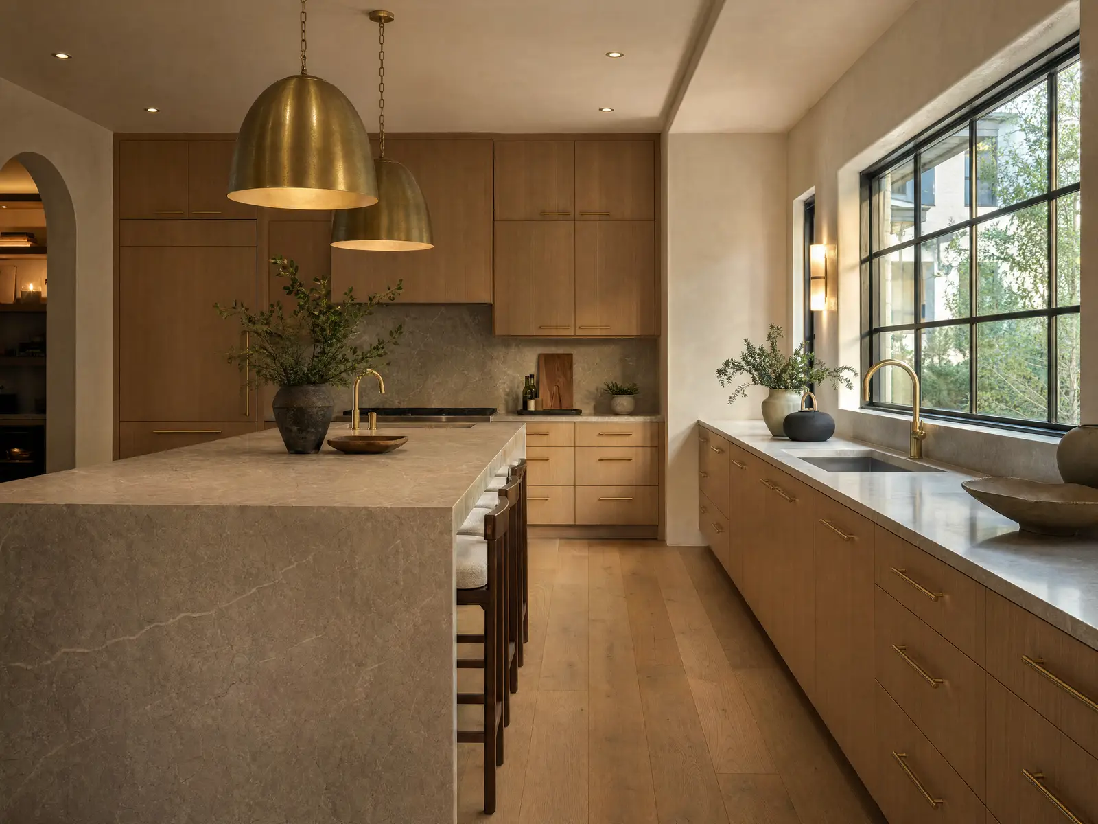 A kitchen at golden hour with full-height white oak cabinetry, a honed stone island with a waterfall edge, and brass pendants over a black steel window.