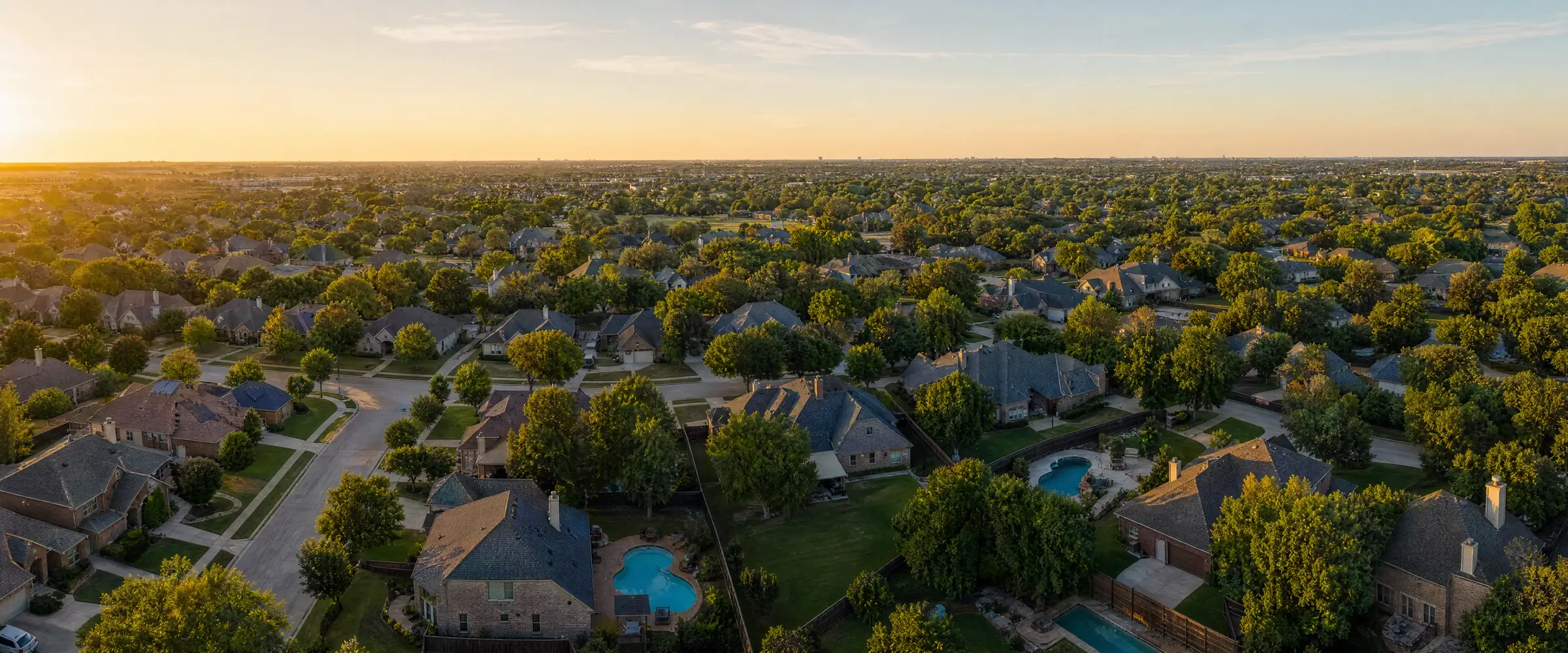 An aerial view of a North Texas residential neighborhood at golden hour, with tree-lined streets, varied rooflines and open sky.